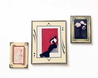 May include: Three framed art prints hang on a white wall. The largest print features a woman in a black dress against a red background. The other two prints have floral and figurative designs, all in decorative frames.