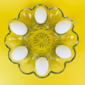 Puede incluir: Un porta huevos de vidrio transparente con seis huevos blancos dentro, sobre un fondo amarillo.