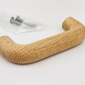 May include: A light-coloured wooden door handle with a curved design. The handle has a textured surface and is accompanied by two screws in a clear plastic bag. The handle is approximately 13 cm long.