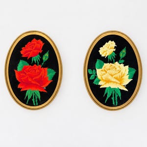 Könnte beinhalten: Zwei ovale Wandbilder mit gestickten Rosenmotiven auf schwarzem Stoff. Ein Bild zeigt leuchtend rote Rosen mit grünen Blättern, das andere hellgelbe Rosen mit grünen Blättern. Beide sind in goldfarbene Rahmen eingefasst.