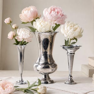 May include: Three silver-toned metal vases of varying heights and shapes, each holding fresh flowers. The center vase is wide and bulbous, while the two flanking vases are slender and trumpet-shaped. Pale pink and white peony blossoms are arranged inside.
