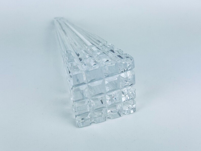 May include: Clear, faceted crystal skewers, arranged in a fan shape. The skewers have a square, geometric design on the handle. The crystal reflects light, creating a sparkling effect. The skewers are on a white background.
