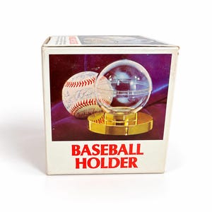 May include: A white cardboard box for a baseball holder featuring a graphic of a clear spherical display case with a gold-coloured base and a signed baseball. The text BASEBALL HOLDER is printed in large red capital letters on the front of the box.