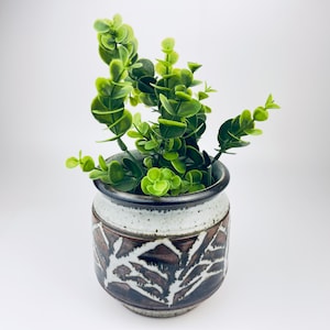 May include: A ceramic plant pot with a speckled white and brown exterior, featuring a dark brown leaf pattern. The pot holds vibrant green faux eucalyptus. The rim is dark brown. A decorative piece for home decor.