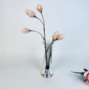 May include: A silver vase with a flared base contains several stems of pink and white artificial flowers. The vase has a slender, curved shape and a decorative design. A few pink roses are in the background.