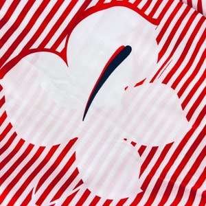 May include: A white fabric featuring a large, stylized flower design. The flower is outlined with red and white stripes. The flower's centre has a dark blue and red accent.