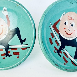 Peut inclure: Deux plateaux en métal de couleur turquoise vintage avec une illustration de dessin animé d'Humpty Dumpty. Le personnage en forme d'œuf est représenté avec un visage souriant, un pantalon noir et un gant blanc, sur fond rouge et blanc.