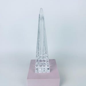 May include: A clear, glass obelisk sculpture with a pyramid shape. The sculpture has vertical lines and sits on a square, light pink base. The background is a plain white.