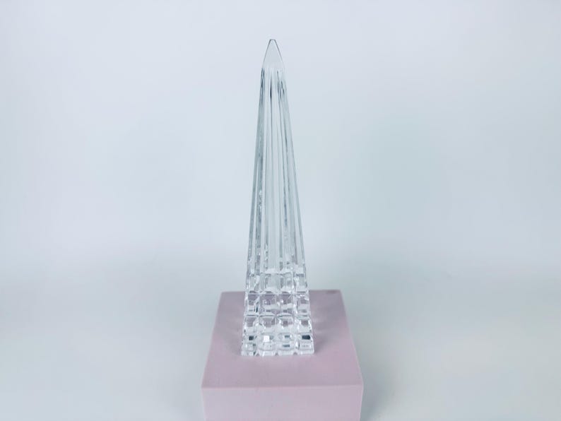 May include: A clear, glass obelisk sculpture with a pyramid shape. The sculpture has vertical lines and sits on a square, light pink base. The background is a plain white.
