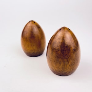 May include: Two wooden salt and pepper shakers. The shakers are egg-shaped with a dark brown finish. Each shaker has three small holes on top. The shakers are on a white background.