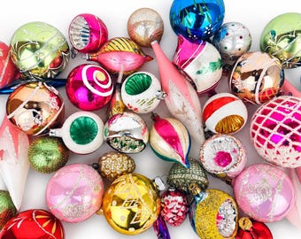 Op de afbeelding: Een verzameling diverse glazen kerstballen in verschillende vormen en maten, met levendige kleuren zoals roze, goud, groen, blauw en zilver, voorzien van reflecterende, glinsterende en gedetailleerde afwerkingen door de hele groep.