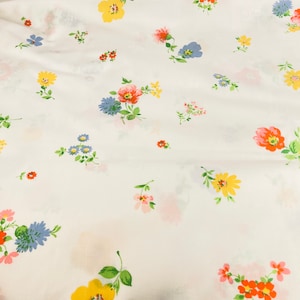 Op de afbeelding: Witte stof met een herhalend patroon van kleine, kleurrijke bloemen. De bloemen zijn in tinten roze, rood, geel en blauw.