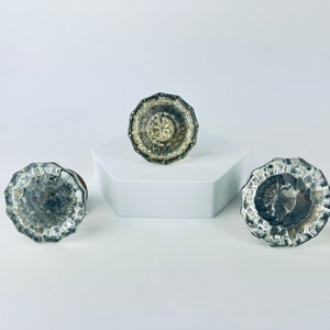 Peut inclure: Trois boutons de porte en verre antique avec une finition argentée et un motif décoratif. Les boutons sont sur une surface blanche.
