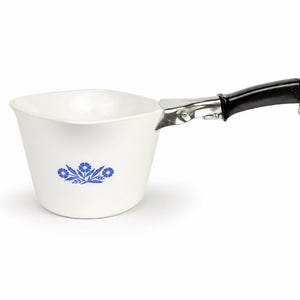 Puede incluir: Un cazo blanco de forma cuadrada con esquinas redondeadas y un estampado floral azul en la parte frontal. Cuenta con un mango negro desmontable con un punto de unión de metal plateado y una pequeña anilla metálica en el extremo para colgarlo.