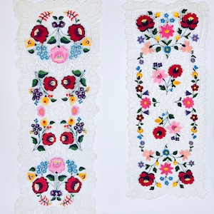 Può includere: Due runner da tavolo bianchi ricamati con disegni floreali colorati. I disegni presentano fiori rossi, rosa, blu e gialli con foglie verdi. I bordi dei runner hanno un delicato bordo in pizzo.