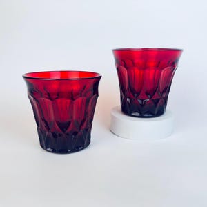 May include: Two ruby red drinking glasses with a faceted design. The glasses have a thick base and a flared rim. One glass sits on a white pedestal. The glasses are made of a transparent material and are likely used for beverages.