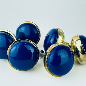 Puede incluir: Cuatro pomos de armario de vidrio azul con ribete de metal dorado. Los pomos son redondos y tienen un acabado liso y brillante.