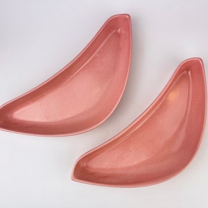 Könnte beinhalten: Zwei rosa, halbmondförmige Servierschalen. Die Schalen haben eine glatte, leicht gesprenkelte Oberfläche und ein flaches, geschwungenes Design. Die Schalen sind wahrscheinlich aus Keramik oder einem ähnlichen Material.