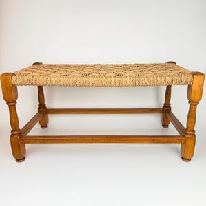 May include: A wooden bench with a woven seat. The bench has four sturdy legs and a rectangular seat made of woven natural fibres. The wood has a warm, light brown finish. The bench is designed for seating.