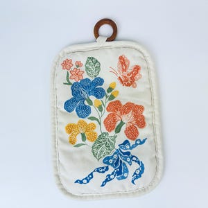 Könnte beinhalten: Ein weißer Topflappen mit einem Blumendesign. Das Design zeigt blaue, orange, gelbe und grüne Blumen und einen Schmetterling.