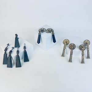 May include: A selection of decorative hardware, including drawer pulls and knobs. The hardware features various designs, including dark bronze-coloured pulls with black tassels, and ornate gold-toned knobs with starburst details and tassels.