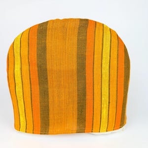 May include: A tea cosy featuring vertical stripes in shades of yellow, orange, and brown. The cosy has a rounded top and a white base. The fabric appears to be woven.
