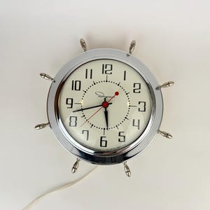 Puede incluir: Un reloj con borde cromado, esfera blanca y números negros. El reloj tiene un diseño de timón de barco con ocho radios plateados. Las manecillas del reloj son negras y rojas. El reloj funciona con un cable blanco.