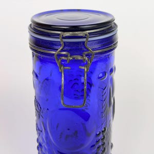 Peut inclure: Un bocal en verre bleu cobalt avec un couvercle et un fermoir en métal. Le bocal présente une surface texturée avec des motifs de raisins et de feuilles en relief. Le couvercle est également bleu.