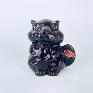 May include: A brown ceramic squirrel ornament with a glossy finish. The squirrel is depicted in a sitting pose, holding its paws together. A red and white sticker is attached. This decorative figurine is a charming item.