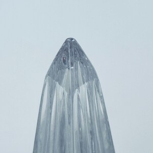 May include: A clear, glass object with a pointed top and vertical, ridged sides. The object is transparent, allowing light to pass through, and is set against a plain, light background. The design suggests a decorative or functional item.