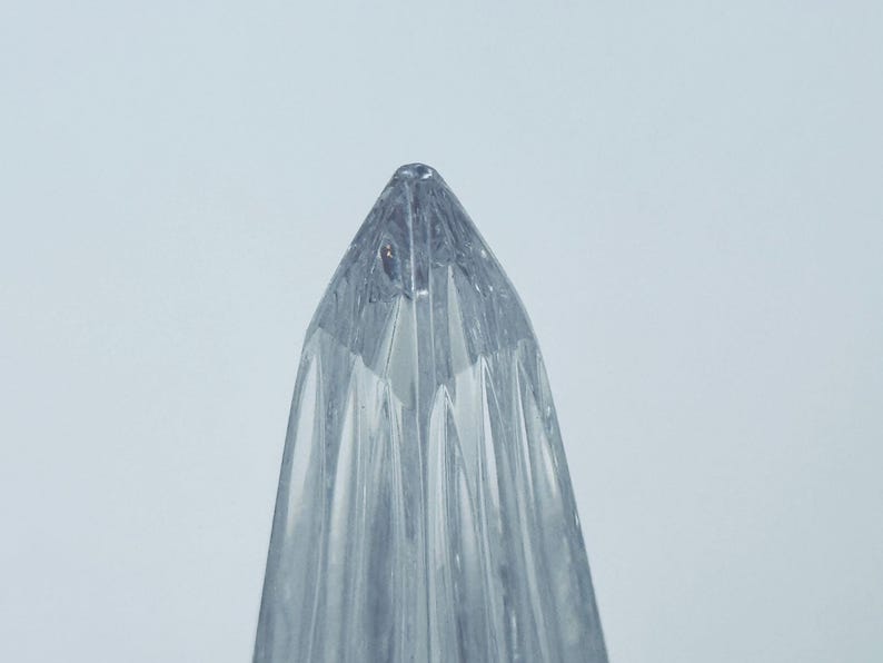 May include: A clear, glass object with a pointed top and vertical, ridged sides. The object is transparent, allowing light to pass through, and is set against a plain, light background. The design suggests a decorative or functional item.