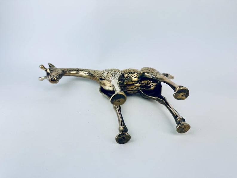 May include: A silver-colored giraffe figurine with detailed etched patterns on its body. The giraffe is lying down, with its legs extended and head turned to the side. The figurine has a detailed design and is made of metal.