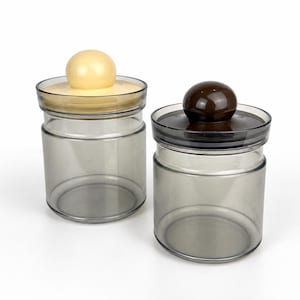 May include: Two cylindrical storage jars made of translucent gray glass. Each jar features a flat lid with a prominent spherical knob handle. One lid is cream-colored, while the other lid is dark brown. The jars have a smooth, clear finish.