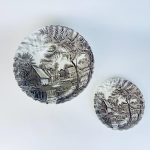 May include: Two decorative plates with a sepia-toned landscape design. The plates feature a detailed illustration of a rural scene with buildings, trees, and a watermill. The plates are round and have a vintage aesthetic.