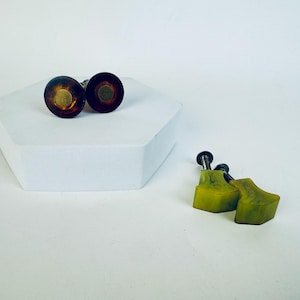 Könnte beinhalten: Zwei Paar dekorative Schrankknöpfe. Ein Paar hat ein dunkelbraunes und goldenes kreisförmiges Design. Das andere Paar hat eine hellgrüne, geometrische Form. Die Knöpfe befinden sich auf einer weißen Oberfläche.