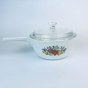 Może przedstawiać: Biały ceramiczny rondel z pokrywką szklaną i wzorem kwiatowym z czerwonymi, żółtymi i brązowymi owocami oraz zielonymi liśćmi.