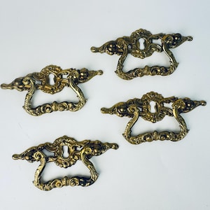 May include: Four antique brass drawer pulls with ornate, scrollwork designs. Each pull features a keyhole and a curved handle. The pulls have a warm, golden hue and are set against a white background.