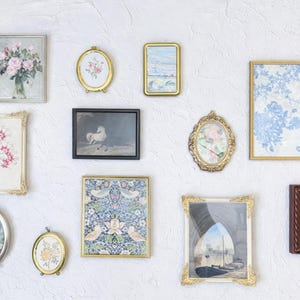 May include: A collection of framed art pieces in various shapes and sizes, including oval, rectangular, and square. The artwork features floral, landscape, and abstract designs, with gold, silver, and brown frames. The pieces are mounted on a textured white wall.