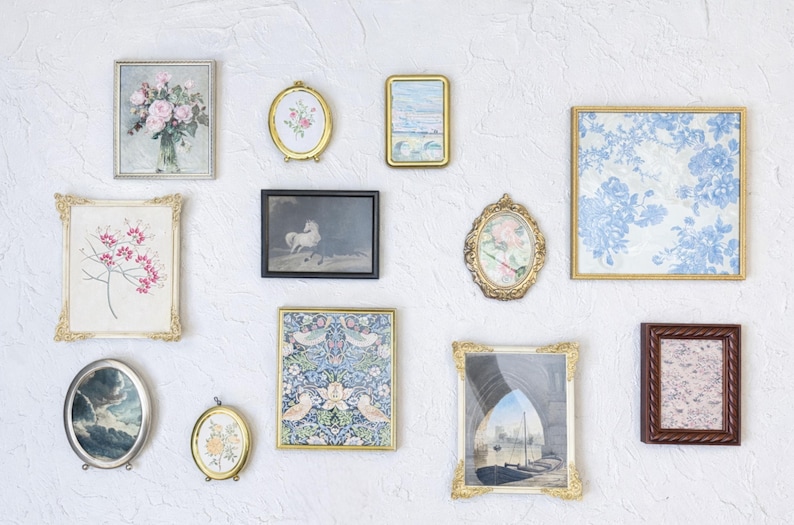 May include: A collection of framed art pieces in various shapes and sizes, including oval, rectangular, and square. The artwork features floral, landscape, and abstract designs, with gold, silver, and brown frames. The pieces are mounted on a textured white wall.