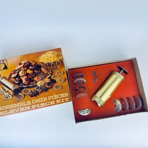 Op de afbeelding: Een vintage koekjespersset met elf onderdelen. De set bevat een gouden koekjespers en een verscheidenheid aan metalen koekjesvormen. De doos is bruin met een afbeelding van koekjes en de tekst "Ensemble Onze Pièces Eleven Piece Kit".