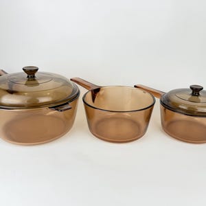 May include: Three amber-colored glass cookware pieces. The set includes a large pot with a lid, a saucepan, and a smaller pot with a lid. The cookware has a classic design with a transparent, heat-resistant construction.