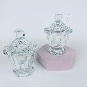 Könnte beinhalten: Zwei klare Glasbehälter mit Deckeln. Ein Behälter hat einen Löffel im Inneren. Die Behälter haben ein dekoratives, facettiertes Design. Die Behälter stehen auf einer hellrosa sechseckigen Oberfläche. Der Hintergrund ist weiß.