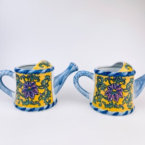 Puede incluir: Dos pequeñas regaderas de cerámica azul y amarilla con diseños florales. Las regaderas tienen un asa y un pico azules.
