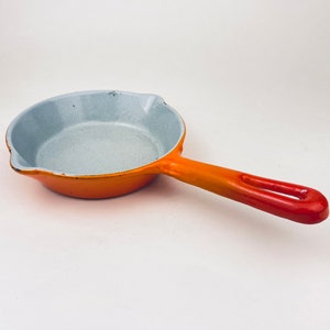 Pode incluir: Frigideira de ferro fundido esmaltada laranja e branca com uma única pega. A frigideira é pequena e tem um interior cinza claro.