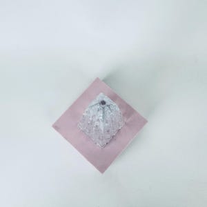 May include: A clear, faceted glass object with a geometric design sits atop a pink square. The glass piece has a diamond-like shape with multiple facets, reflecting light. The background is a plain white surface.