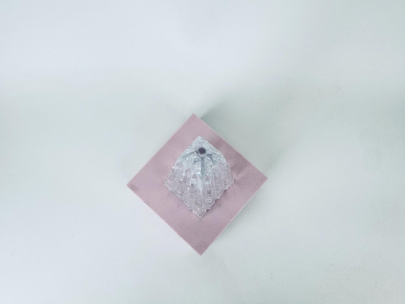 May include: A clear, faceted glass object with a geometric design sits atop a pink square. The glass piece has a diamond-like shape with multiple facets, reflecting light. The background is a plain white surface.