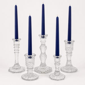 May include: A set of five clear glass candle holders of varying heights and designs, each featuring a tall, slender, solid navy blue taper candle. The glass holders have intricate pressed patterns, faceted bases, and textured stems.