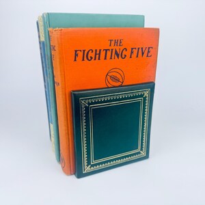 May include: A stack of vintage books with colorful covers. The book in front has an orange cover with the title "THE FIGHTING FIVE" in black lettering. A dark green bookend with gold trim is in front.