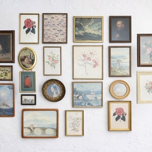 May include: A collection of framed vintage art pieces, including floral prints, landscapes, and portraits, arranged on a white wall. Frames vary in size, shape, and color, with gold, brown, and black frames. The art pieces add a touch of classic decor.