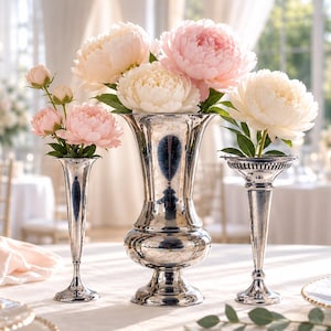 May include: A set of three silver-toned metal vases with a polished, reflective finish. The central vase is large with a wide, flared rim and a rounded base. Two smaller, slender trumpet-shaped vases flank the center, each holding pink and white peonies.